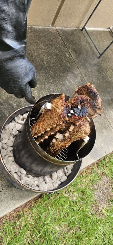 Gallery 105 Close-up of meat grilling on a barbecue with a gloved hand in an outdoor setting.