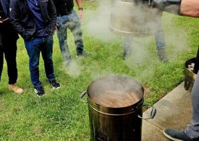 Gallery 101 People gathered around a steaming outdoor cooking pot on a grassy area.