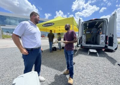 Gallery 24 Two men conversing outdoors near a yellow canopy and mobile testing van under a blue sky.