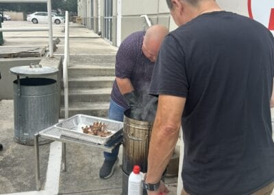 Gallery 44 Two men preparing food outdoors near a warehouse with a trash can and parking lot in the background.