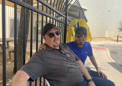 Gallery 46 Two men sitting outdoors near a black metal fence, wearing sunglasses and casual clothing.